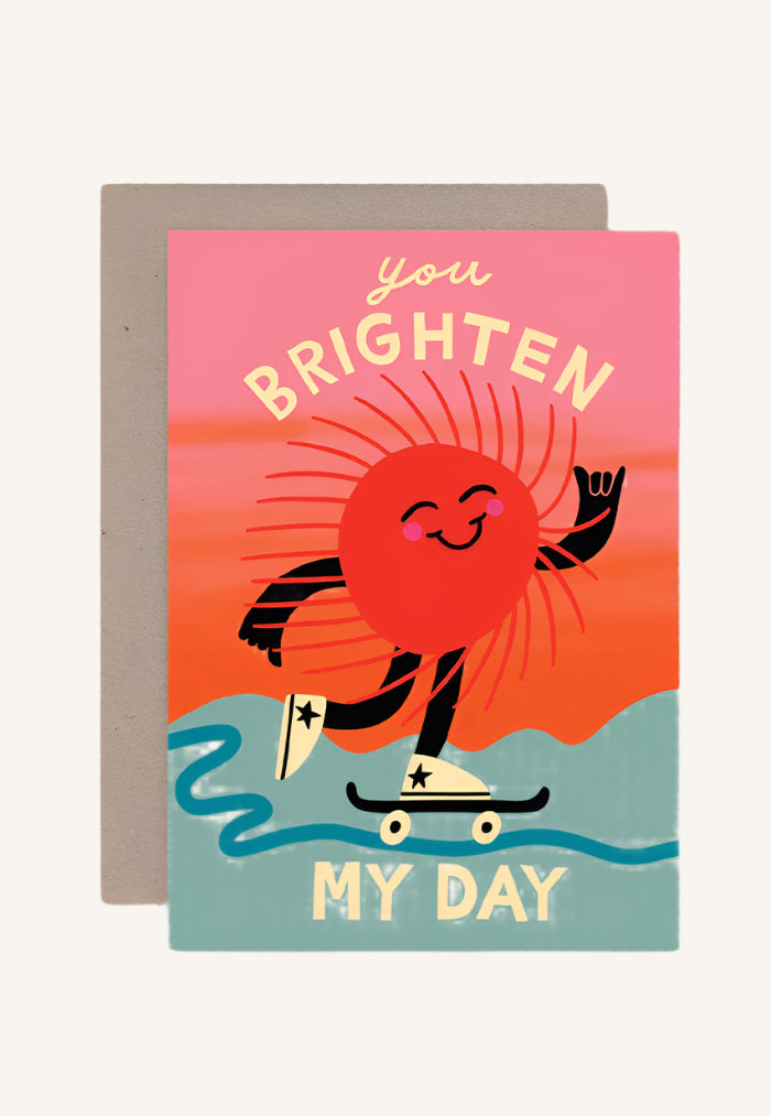 Greeting card with illustration of a sun character on a skateboard with text 'you BRIGHTEN MY DAY' on a pink background. Includes a brown envelope.
