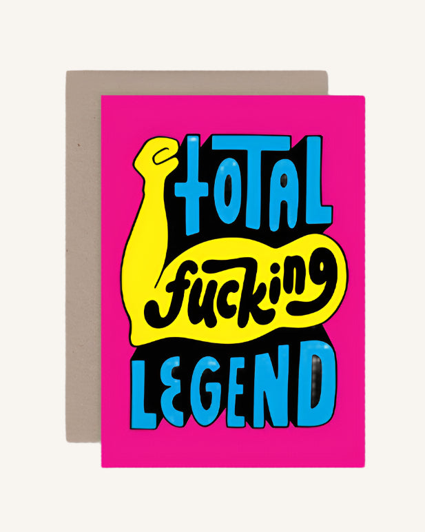 A greeting card with bold text 'TOTAL FUCKING LEGEND' in yellow and blue on a pink background, with a brown envelope.