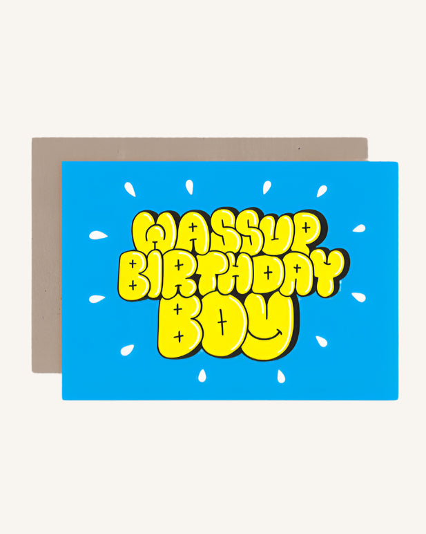 A blue greeting card with the text 'WASSUP BIRTHDAY BOY' in bold yellow letters, accompanied by a brown kraft envelope.